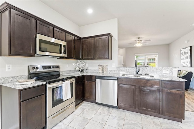 kitchen featuring stainless steel appliances, a sink, a peninsula, light stone counters, and dark brown cabinetry