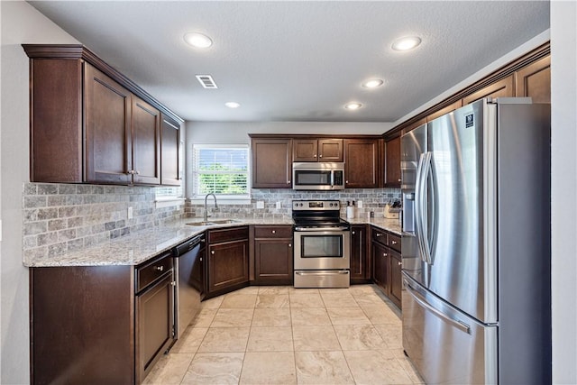 kitchen with appliances with stainless steel finishes, a sink, dark brown cabinets, decorative backsplash, and light stone counters