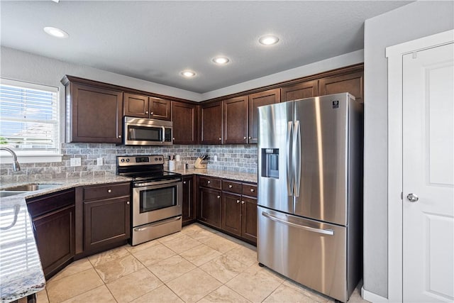 kitchen featuring stainless steel appliances, a sink, dark brown cabinetry, backsplash, and light stone counters