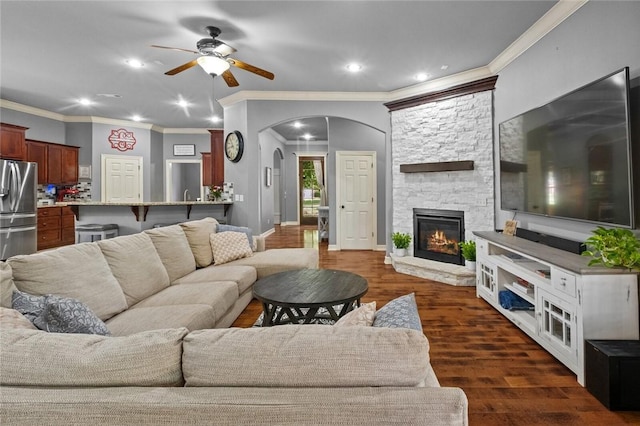 living area with ceiling fan, crown molding, dark wood-style floors, arched walkways, and a stone fireplace