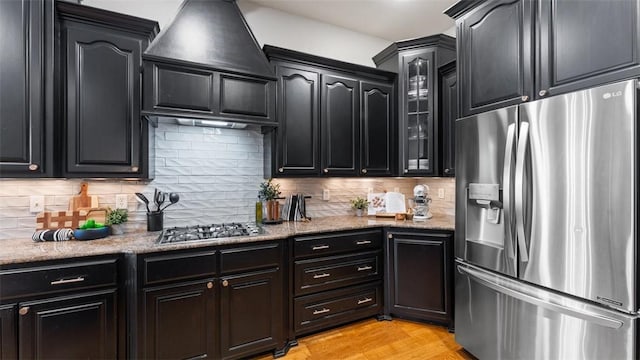 4903 S Inglewood Rd, Rogers, AR 72758 - kitchen with appliances with stainless steel finishes, custom range hood, dark cabinetry, glass insert cabinets, and backsplash