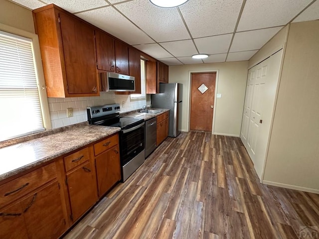 kitchen with brown cabinets, backsplash, appliances with stainless steel finishes, dark wood-type flooring, and baseboards