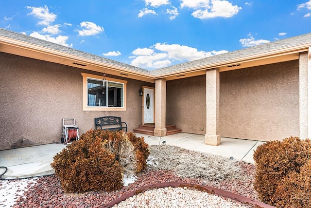 view of exterior entry with a shingled roof, a patio, and stucco siding