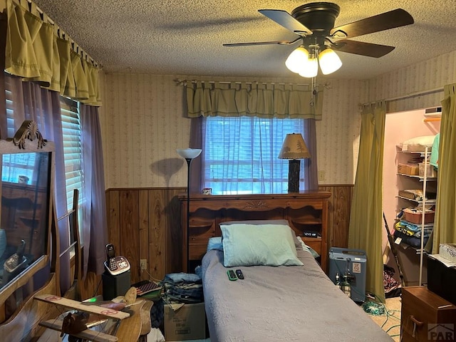 bedroom featuring a ceiling fan, a wainscoted wall, a textured ceiling, and wallpapered walls