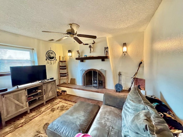 living room with a textured ceiling, a fireplace with raised hearth, ceiling fan, a textured wall, and light wood-style flooring