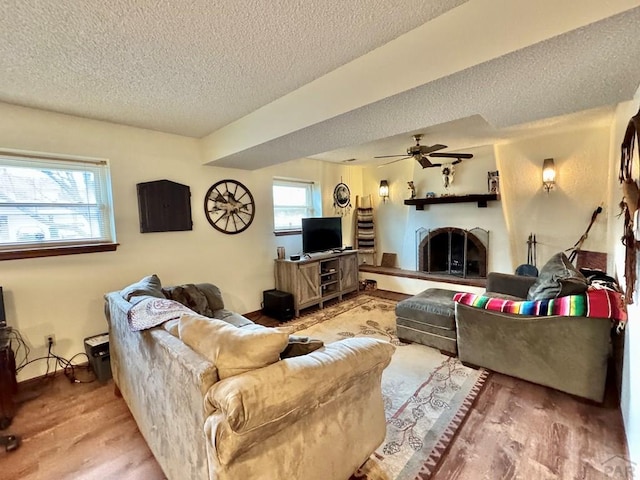 living room featuring light wood-type flooring, a ceiling fan, a fireplace with raised hearth, and a textured ceiling