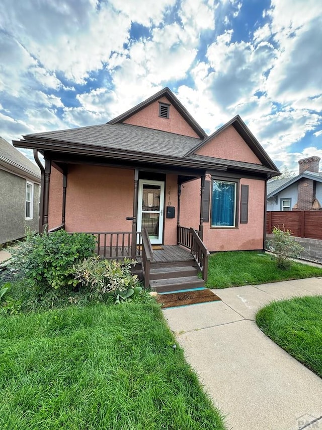 bungalow-style home featuring a shingled roof, covered porch, fence, a front lawn, and stucco siding