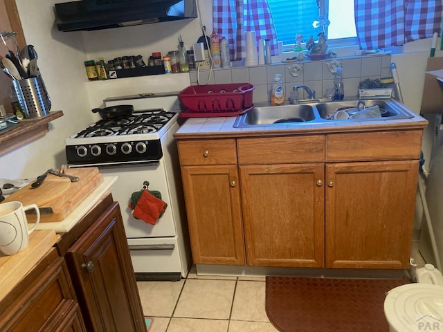 kitchen featuring a sink, under cabinet range hood, brown cabinets, tile countertops, and white gas range oven