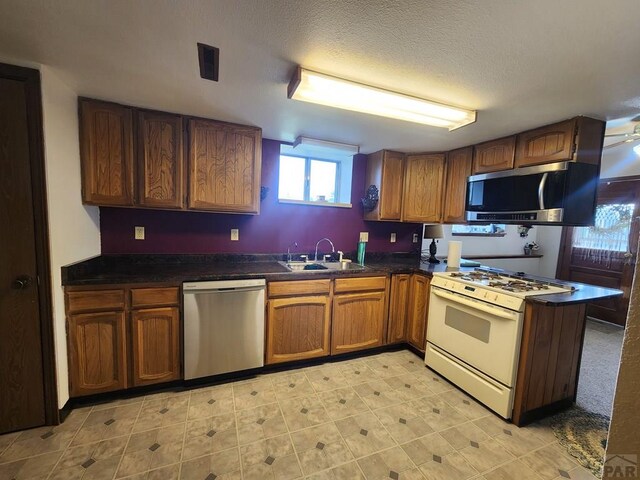 kitchen with a textured ceiling, brown cabinets, a sink, dark countertops, and stainless steel appliances