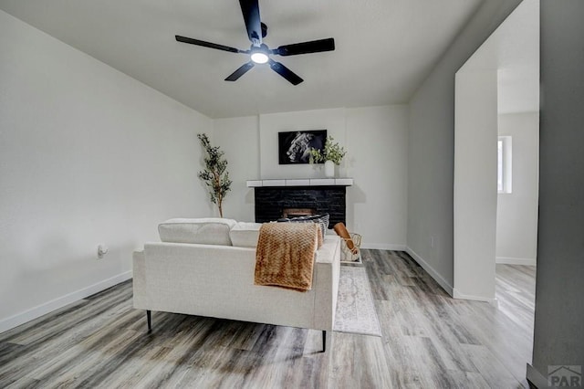 living room featuring ceiling fan, a stone fireplace, light wood-type flooring, and baseboards