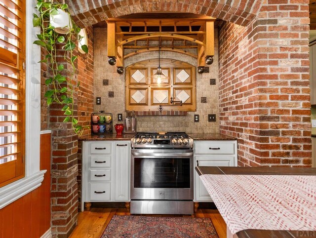 kitchen featuring stainless steel gas stove, white cabinetry, brick wall, wood finished floors, and dark stone counters