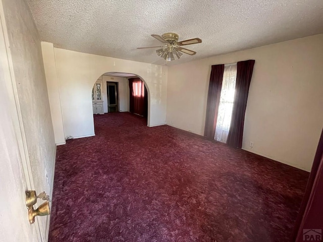 unfurnished room featuring arched walkways, a textured ceiling, dark carpet, and a ceiling fan