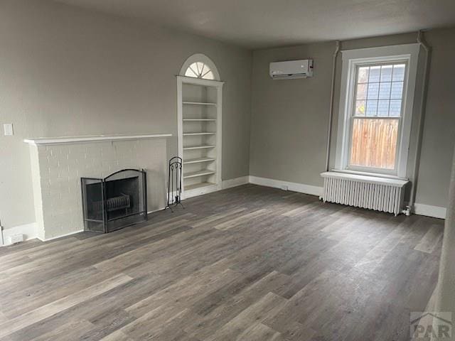 unfurnished living room featuring built in shelves, radiator heating unit, a fireplace with flush hearth, dark wood-type flooring, and an AC wall unit