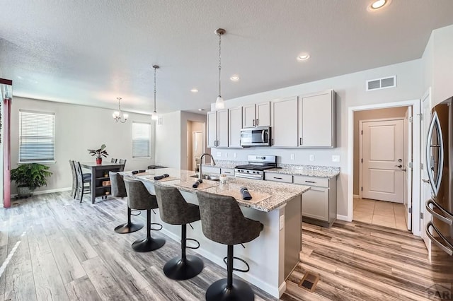 kitchen featuring light stone counters, a center island with sink, gray cabinets, hanging light fixtures, and appliances with stainless steel finishes