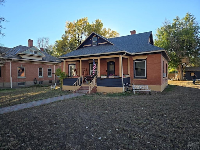 view of front facade featuring brick siding, a chimney, a shingled roof, covered porch, and a front lawn