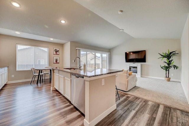 kitchen featuring a center island with sink, dishwasher, a glass covered fireplace, light stone countertops, and a sink