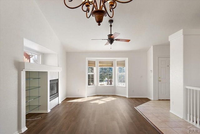 unfurnished living room with a tiled fireplace, ceiling fan with notable chandelier, wood finished floors, and baseboards