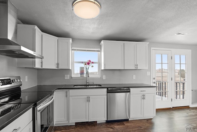 kitchen with a sink, appliances with stainless steel finishes, dark countertops, and wall chimney range hood