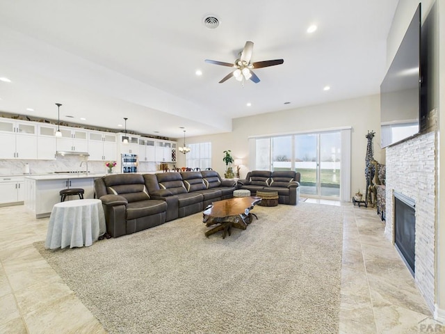 living area featuring plenty of natural light, ceiling fan with notable chandelier, visible vents, and a stone fireplace