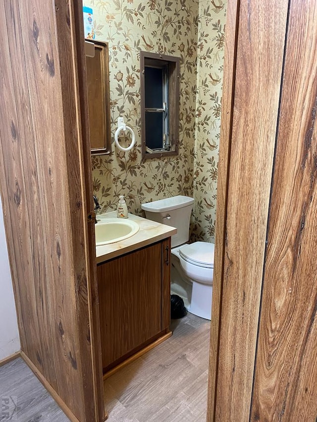 half bathroom with wallpapered walls, vanity, toilet, and wood finished floors