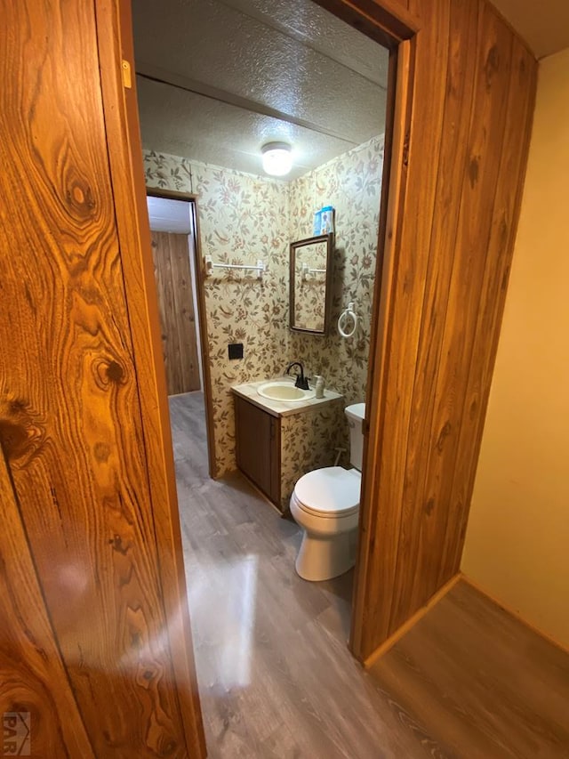 half bathroom with a textured ceiling, toilet, wood finished floors, vanity, and wallpapered walls