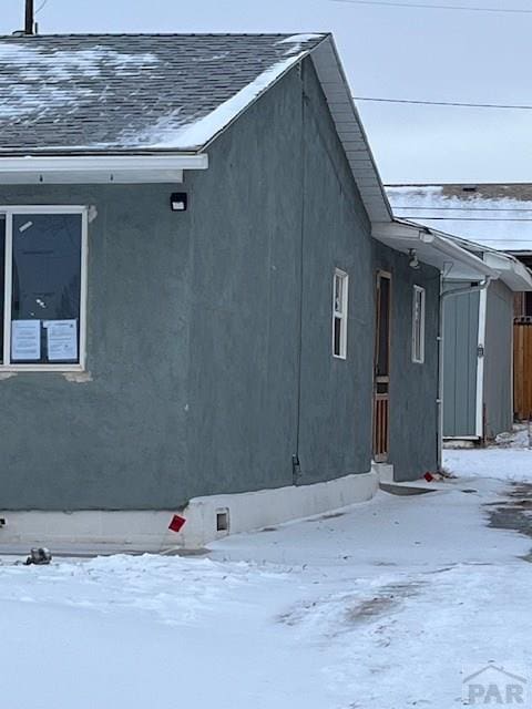 view of snowy exterior featuring stucco siding, fence, and roof with shingles