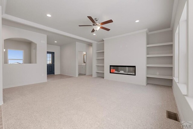 unfurnished living room featuring a ceiling fan, light colored carpet, a glass covered fireplace, crown molding, and built in shelves