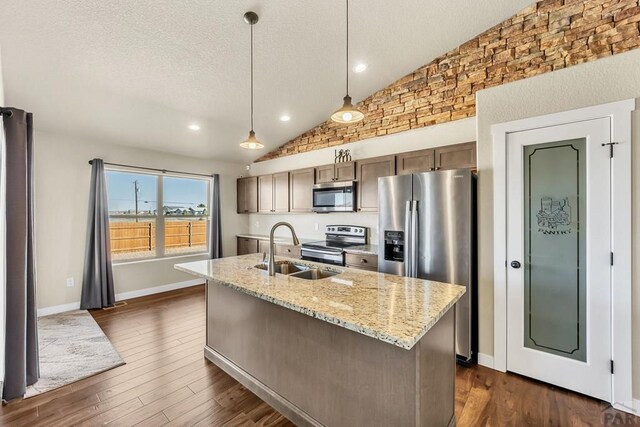 kitchen with light stone counters, a kitchen island with sink, a sink, appliances with stainless steel finishes, and decorative light fixtures