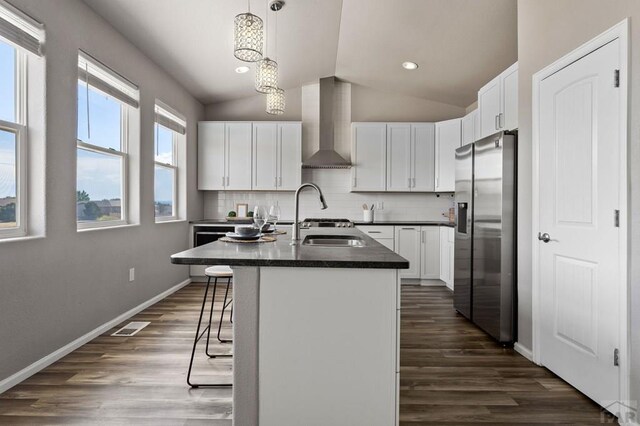 kitchen featuring a breakfast bar, stainless steel refrigerator with ice dispenser, dark countertops, wall chimney range hood, and an island with sink