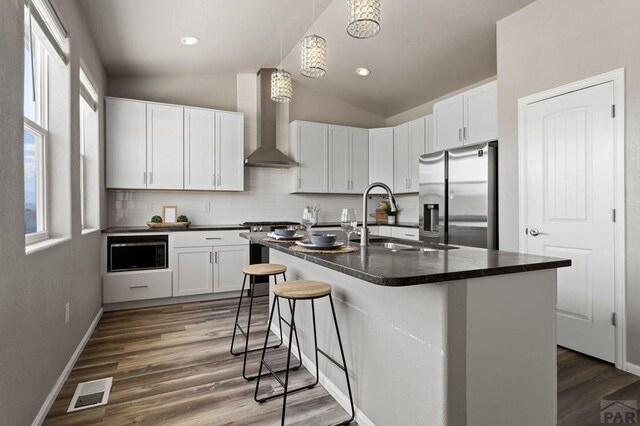 kitchen with white cabinetry, appliances with stainless steel finishes, wall chimney exhaust hood, dark countertops, and a center island with sink