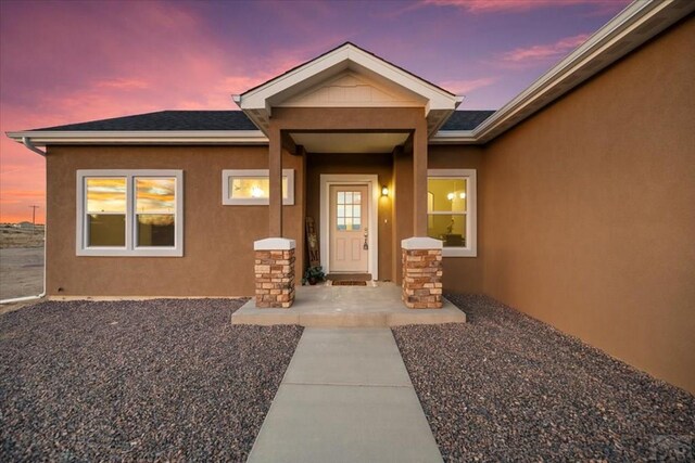 exterior entry at dusk with roof with shingles and stucco siding