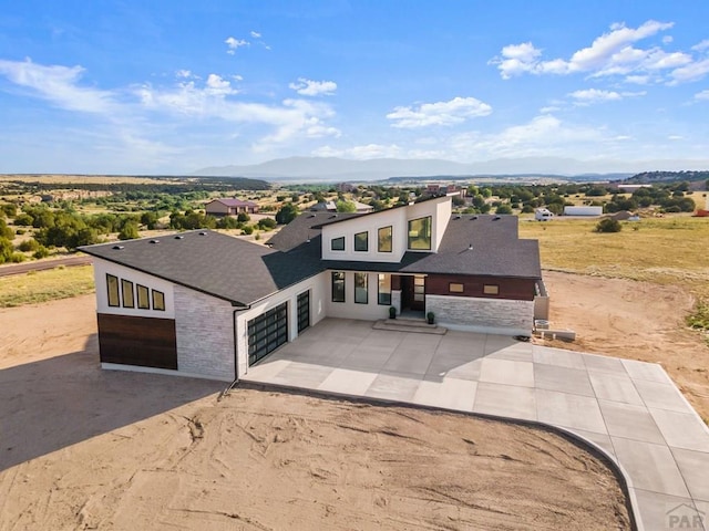 contemporary house with driveway, stone siding, a mountain view, an attached garage, and a shingled roof