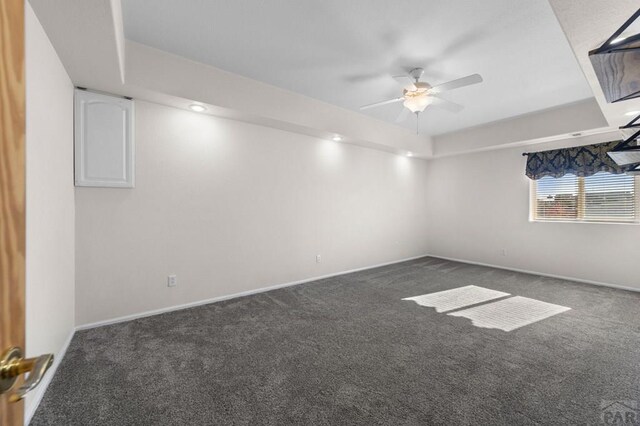 unfurnished room with a tray ceiling, dark carpet, a ceiling fan, and baseboards