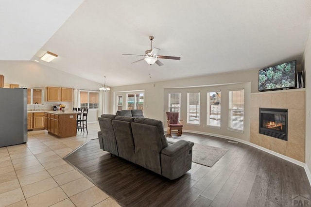 living room featuring light wood finished floors, baseboards, vaulted ceiling, a fireplace, and ceiling fan with notable chandelier