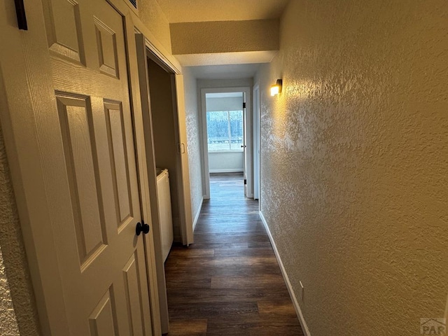 hall with dark wood-style flooring, a textured wall, and baseboards