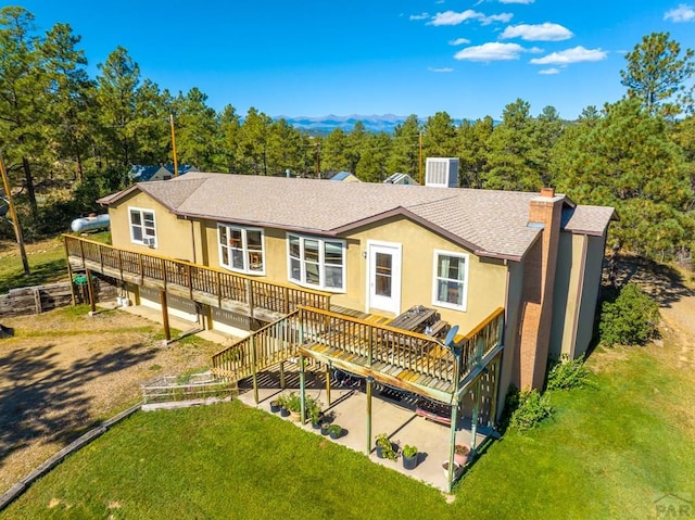 back of house featuring a shingled roof, a lawn, a wooden deck, stucco siding, and a chimney