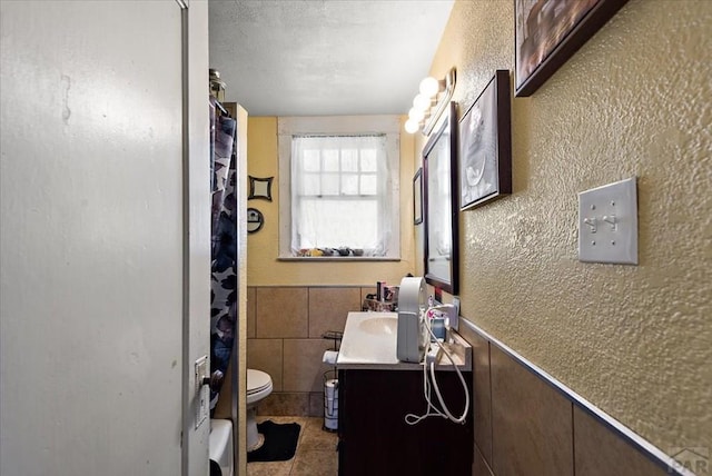 full bathroom featuring a textured wall, toilet, wainscoting, tile patterned flooring, and vanity