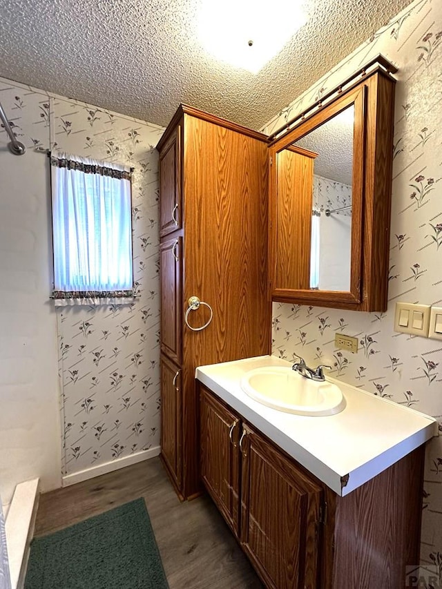 bathroom featuring a textured ceiling, wood finished floors, vanity, baseboards, and wallpapered walls