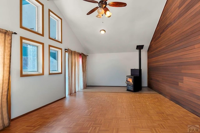 unfurnished living room featuring a wood stove, wood walls, ceiling fan, high vaulted ceiling, and baseboards