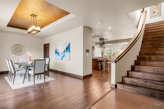 dining space featuring dark wood-style flooring, a notable chandelier, a raised ceiling, stairway, and baseboards