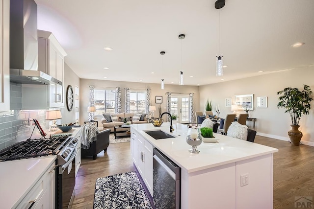 kitchen with light countertops, open floor plan, a sink, wall chimney range hood, and an island with sink