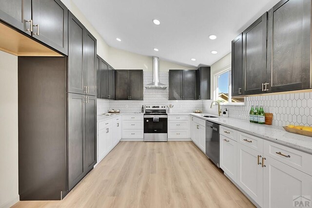 kitchen with vaulted ceiling, stainless steel appliances, a sink, light wood finished floors, and wall chimney exhaust hood