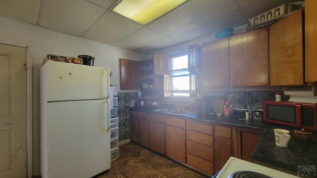 kitchen featuring freestanding refrigerator, brown cabinetry, a drop ceiling, a sink, and dark countertops