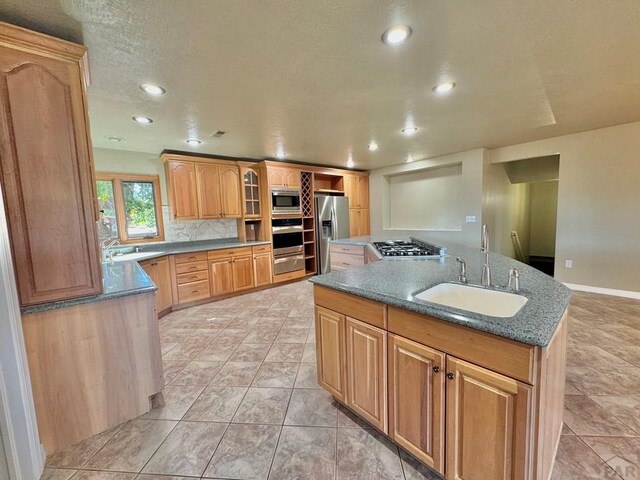 kitchen with a sink, appliances with stainless steel finishes, decorative backsplash, an island with sink, and glass insert cabinets