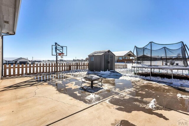 snow covered patio featuring a trampoline, a fenced backyard, an outdoor structure, and a shed