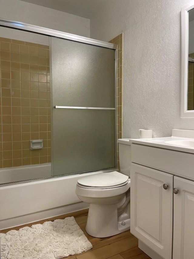 bathroom with bath / shower combo with glass door, a textured wall, toilet, wood finished floors, and vanity