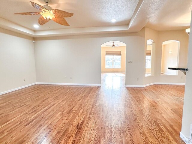 unfurnished room with a tray ceiling, arched walkways, light wood finished floors, a ceiling fan, and baseboards