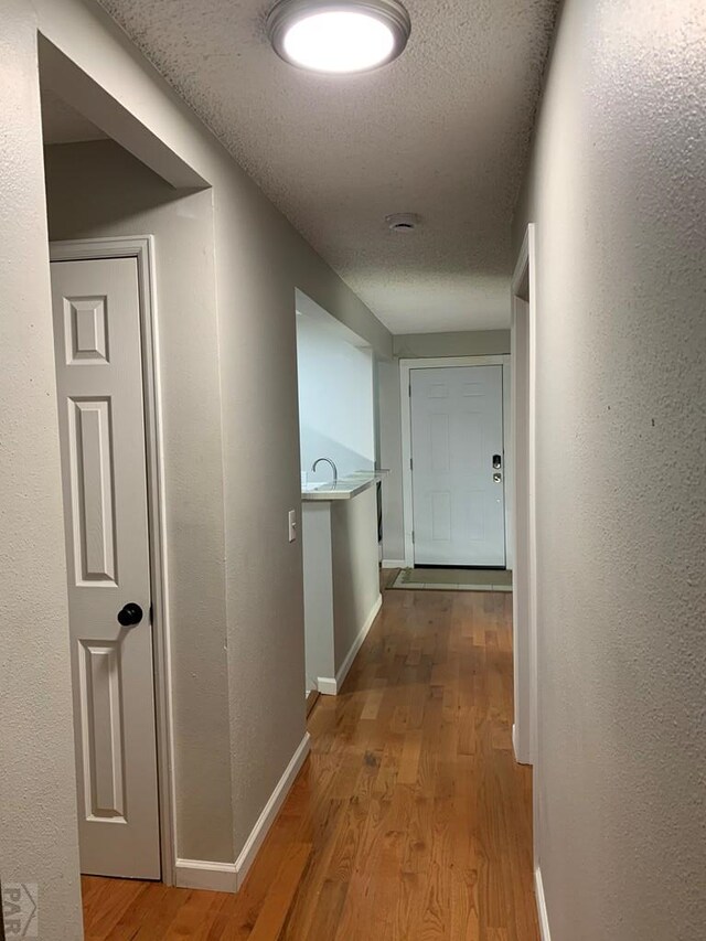 hallway featuring light wood-type flooring, a textured wall, a textured ceiling, and baseboards