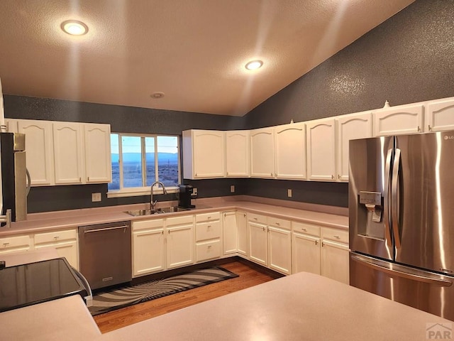 kitchen with a textured wall, stainless steel appliances, a sink, white cabinets, and vaulted ceiling