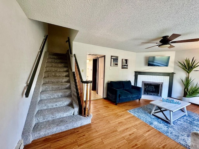 unfurnished living room with a tiled fireplace, ceiling fan, a textured ceiling, wood finished floors, and stairs